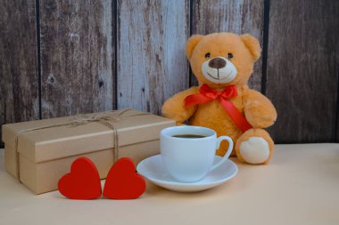 Kahve fincanının yanında oyuncak ayı ve ahşap arka fonda paketlenmiş hediyeler, buhar fincanı, kurdeleli paketler, kırmızı kalp jetonları, samimi bir kahvaltı.