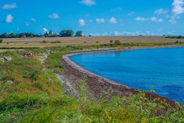 Turkuaz Körfezi ve Çakıl Taşı Sahil Hattı, Sakin Şeffaf Su, Uzak Kıyıdaki Küçük Kulübe Kümesi, Canlı Yeşil Sebze Kıyısı, Parlak Gün ortası