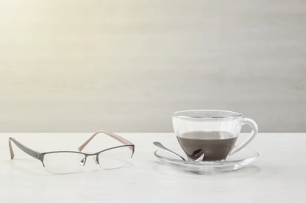 Nahaufnahme schwarzer Kaffee in durchsichtiger Tasse Kaffee und Brille auf verschwommenem Holztisch und Wand strukturiertem Hintergrund, Entspannung im Kaffeepausenkonzept durch Kaffee und Brille im Kunstton — Stockfoto