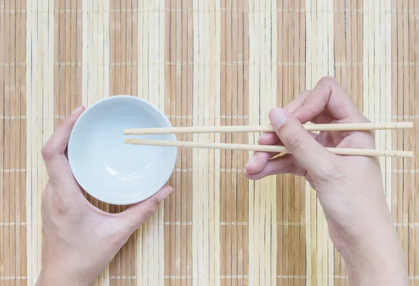 Primer plano cáliz de cerámica blanca con palillos de madera borrosa en la mano de la mujer en la estera de madera fondo texturizado en la mesa de comedor en la vista superior en el concepto de comer — Foto de Stock