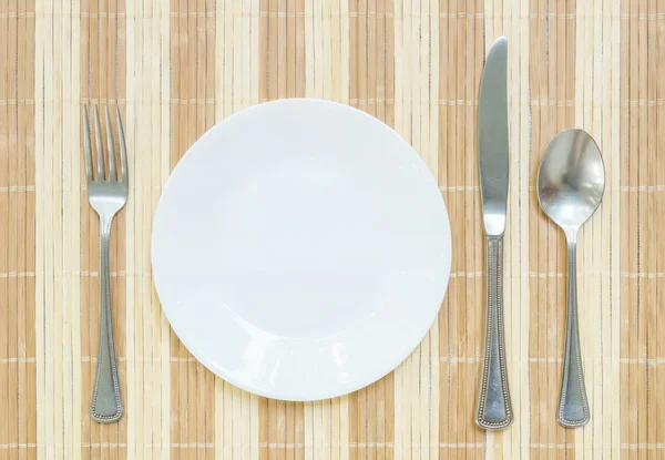 Närbild vitt keramikfat med rostfritt gaffel och sked och kniv på trä matta texturerat bakgrund på matbord i ovanifrån — Stockfoto