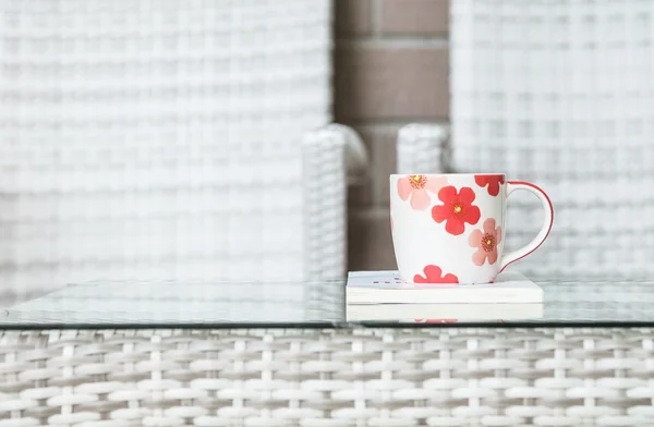 Nahaufnahme niedliche Tasse auf weißem Buch auf verschwommenem Holz weben Tisch und Stuhl strukturierten Hintergrund — Stockfoto