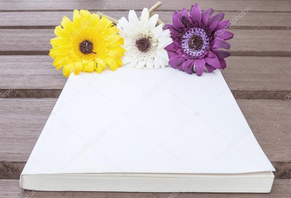 Closeup white note book with colorful faked flower on blurred old wood table textured background