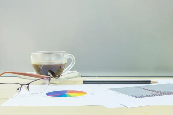 Κοντινό μαύρο καφέ σε διαφανές φλιτζάνι καφέ με χαρτί εργασίας και μολύβι και γυαλιά σε θαμπό ξύλινο γραφείο και παγωμένο γυάλινο τοίχο με υφή φόντο, έννοια εργασίας με καφέ και χαρτί εργασίας — Φωτογραφία Αρχείου