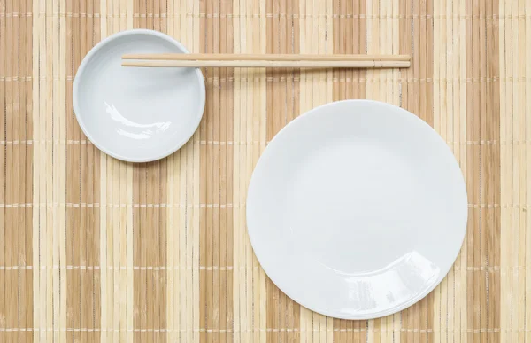 Primer plano plato de cerámica blanca y cáliz con palillos de madera en la estera de madera fondo texturizado en la mesa de comedor en vista superior — Foto de Stock