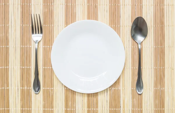 Plato cerámico blanco de primer plano con tenedor inoxidable y cuchara sobre alfombra de madera fondo texturizado sobre mesa de comedor en vista superior — Foto de Stock