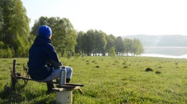 ahşap skomeyke ve dökülen kendini bir termos çaydan oturan bir adam. güneşin doğuşunu gölde izliyor. Göl üzerinde sis. Güneş doğuyor. çay içme ve doğa hayran bir adam