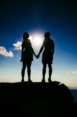 Bir dağın tepesinde el ele tutuşan sevgi dolu bir çift. Bir erkek ve bir kız birbirlerine dik dik bakarlar.