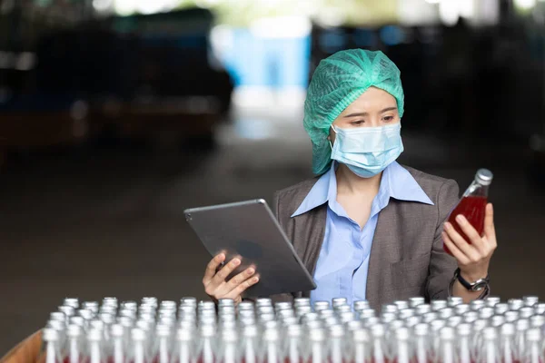 young woman manager wearing safety hat and medical mask for protect virus(covid-19) using tablet computer for stocktaking of products in beverage factory