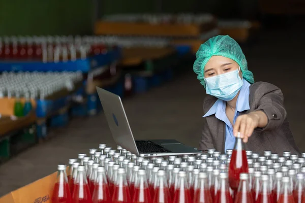 young woman manager wearing safety hat and medical mask for protect virus(covid-19) using laptop computer and picking up beverage for stocktaking of products in beverage factory