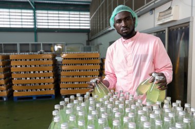 Afrikalı erkek fabrika işçisi meşrubat fabrikasındaki kaliteyi kontrol etmek için yeşil meyve suyu şişesi veya fesleğen tohumu içeceği alıyor.