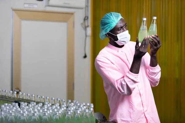 African male factory worker wearing medical mask, checking and looking at green juice bottle or basil seed drink in beverage factory