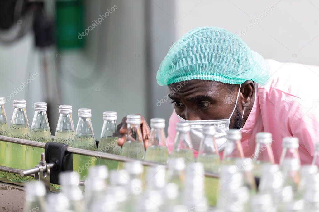 African male factory worker wearing medical mask, picking up green juice bottle or basil seed drink for checking quality in beverage factory