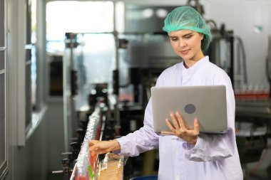 Genç kadın fabrika işçisi fesleğen tohumu içeceği topluyor ve içecek fabrikasının kalitesini kontrol etmek için dizüstü bilgisayar kullanıyor.