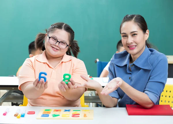 portrait asian disabled child or down syndrome child showing number puzzle and woman teacher helping in classroom