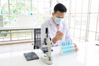 Tıbbi yüz maskesi takan bilim adamı laboratuvarda kimyasal bir deney yapıyor ya da test ediyor.