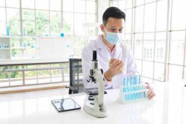 Tıbbi yüz maskesi takan bilim adamı laboratuvarda kimyasal bir deney yapıyor ya da test ediyor.