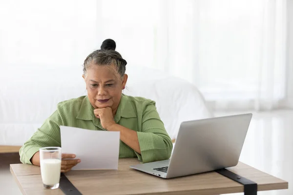 senior woman looking paper document and using laptop computer at home