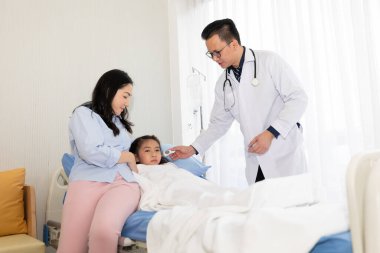 Asyalı doktor çocuğun kulağının ısısını ölçüyor ve annesi hastanede yanındaymış. 