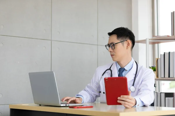 asian doctor talking with patient and co-worker through online video chat with computer and holding tablet in hospital