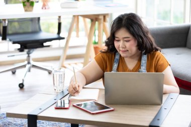 Tombul kadın dizüstü bilgisayara bakıyor ve çalışmak için not defterine yazıyor.
