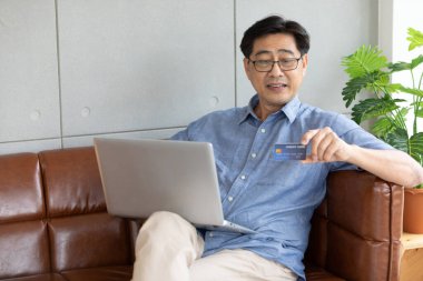 Üst düzey Asyalı adam online alışveriş yapmak ve kredi kartıyla ödeme yapmak için dizüstü bilgisayar kullanıyor.