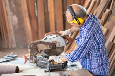Kıdemli Asyalı marangoz gözlük ve kulaklık takıyor, elektrikli testere kullanarak atölyedeki bir tahta parçasında tahta kesiyor.