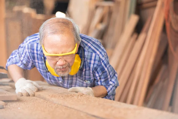 senior asian man carpenter blowing off sawdust from plank of wood in carpentry workshop and he\'s suffering itching scratching eyes