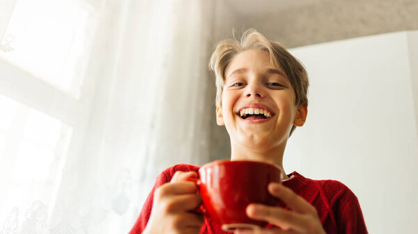  Happy laughing teenager boy holds a red cup with cocoa and marshmallows. Soft selective focus.