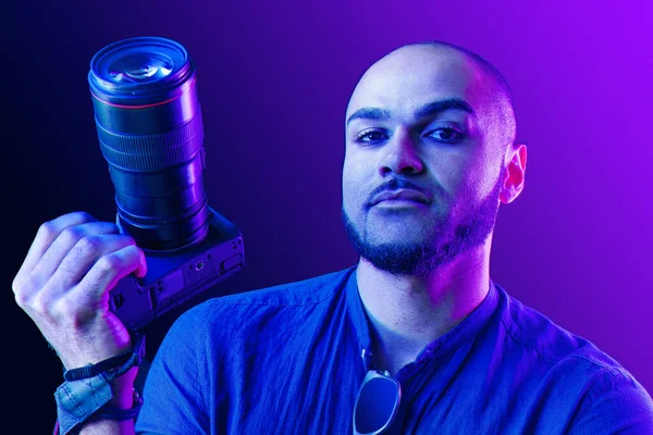 Black man with camera standing against purple background in neon light