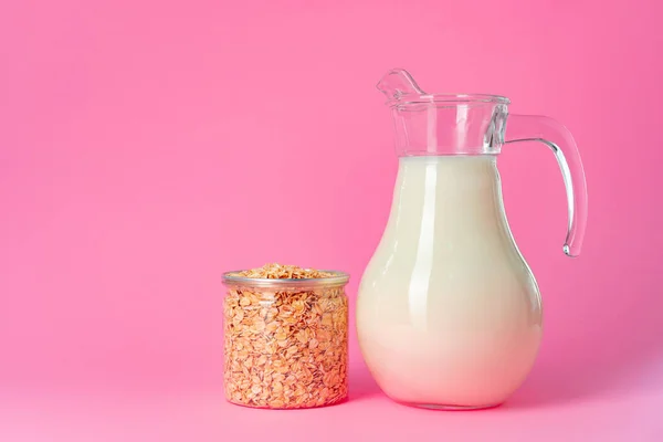 Oat milk in glassware and dry oat flakes on pink background