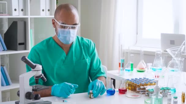Ученый-вирусолог готовится к введению вакцины в шприц из пробирки, сидя за столом в лаборатории — стоковое видео