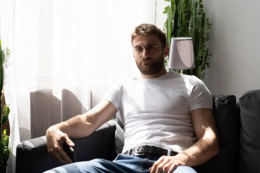 Pencerenin yanındaki koltukta oturan üzgün, düşünceli, sakallı adam.