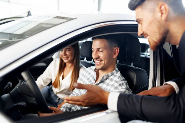 Pareja joven eligiendo un coche en el concesionario con el gerente de ayudar — Foto de Stock