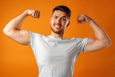 Güçlü kollarını sarı arka planda gösteren pozitif gülümseyen genç adam.