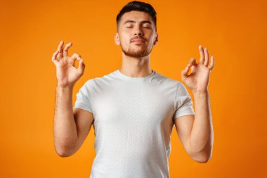 Rahatlamış bir adam, meditasyon yaparken sarı arka planda duruyor.