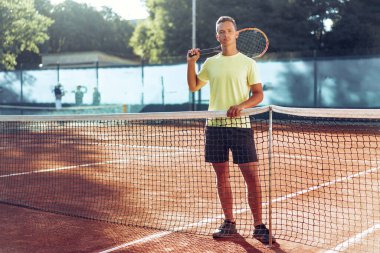 Tenis raketli genç adam kil sahada ağın yanında duruyor.