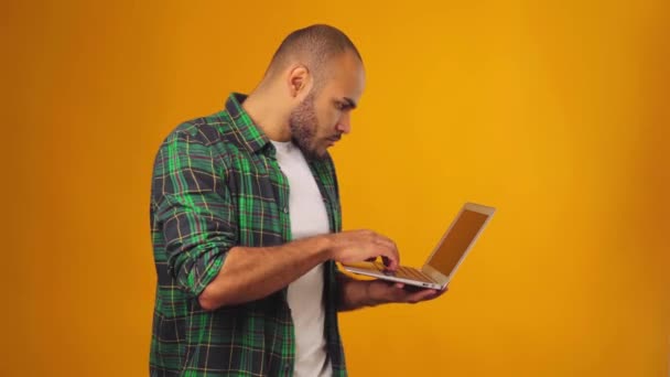 African American young man in green shirt standing against yellow background and typing on laptop computer — Stock Video