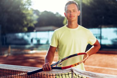 Tenis raketli genç adam kil sahada ağın yanında duruyor.
