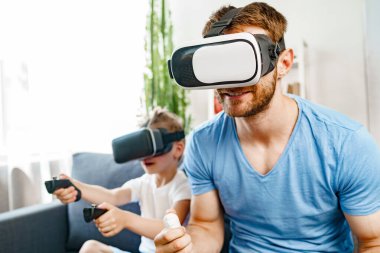 Baba oğul kanepede oturup VR gözlükleriyle oyun oynuyorlar.