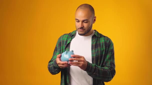 Clueless african american man shaking piggy bank and find out there is no money against yellow background — Stock Video