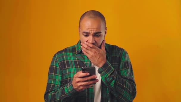 Африканский американец в изумлении смотрит на экран смартфона и робко закрывает лицо. — стоковое видео
