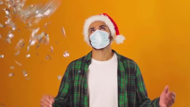Hombre triste en Santa sombrero y máscara médica de pie bajo la caída de confeti contra el fondo amarillo — Vídeos de Stock