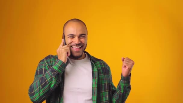 Hombre afroamericano positivo hablando por teléfono y sonriendo de buenas noticias con el pulgar hacia arriba sobre el fondo amarillo — Vídeos de Stock