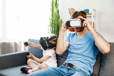 Baba oğul kanepede oturup VR gözlükleriyle oyun oynuyorlar.