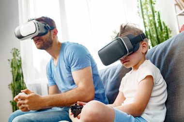 Baba oğul kanepede oturup VR gözlükleriyle oyun oynuyorlar.