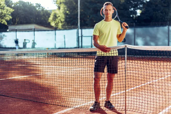 Tenis raketli genç adam kil sahada ağın yanında duruyor. — Stok fotoğraf