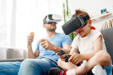 Baba oğul kanepede oturup VR gözlükleriyle oyun oynuyorlar.