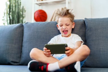 Evdeki kanepede oturan ve akıllı telefon kullanan küçük çocuk.