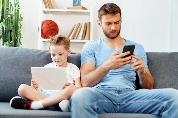 Маленький хлопчик використовує цифровий планшет і його батько, використовуючи смартфон, сидить разом на дивані — стокове фото